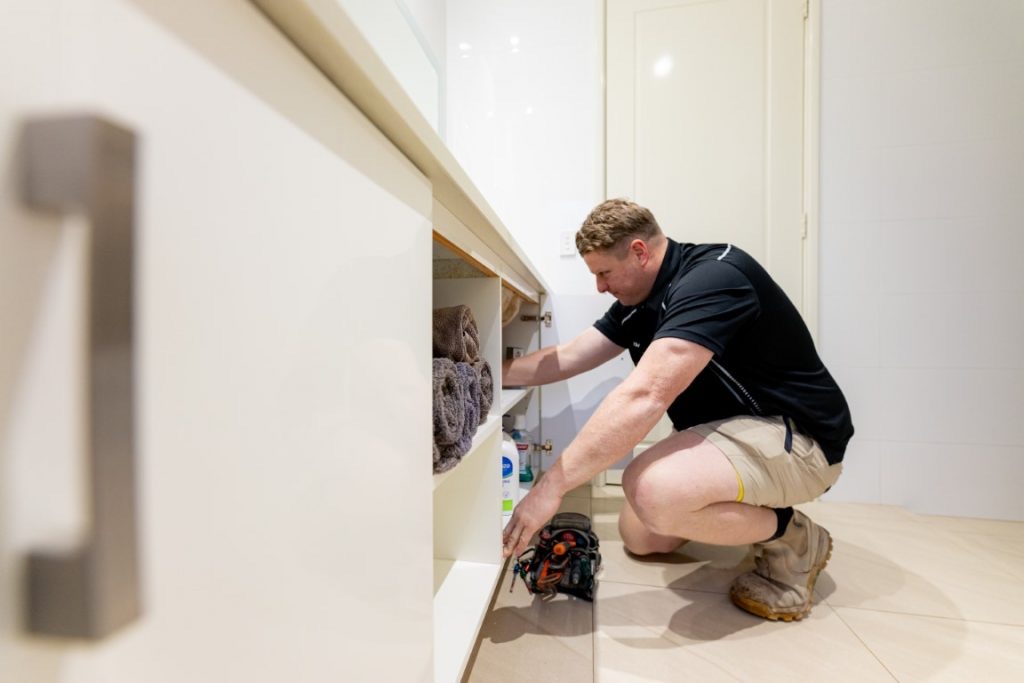 Plumber crouched at an open vanity cupboard with a tool pouch beside him on the tiles
